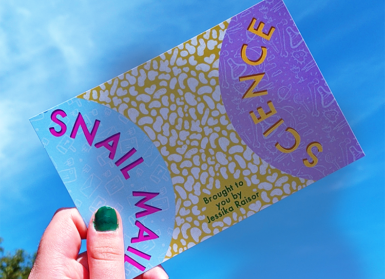 A postcard that says "snail mail science" being held up against a blue sky