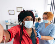 Self portrait of Black woman with protective face mask shooting selfie in the doctor's office while getting a COVID-19 vaccine